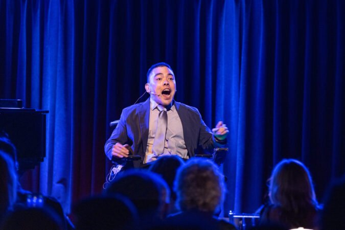 BRENNAN SRISIRIKUL is a Asian American 30 something with youthful looks in a suit and tie singing in front of a blue curtain.