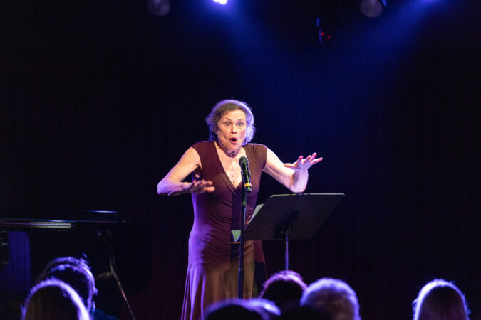 ANITA HALLENDER singing into a microhone on a stand with a wildly expression on her face. Anita is an older white woman wearing a purple dress.