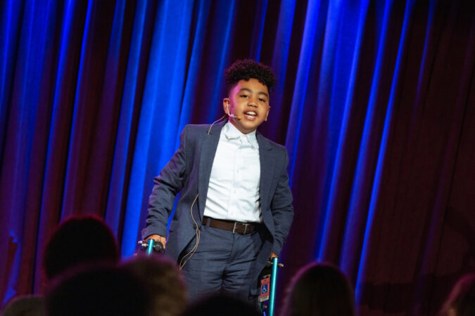 SEBASTIAN ORTIZ is a Afro-Latinx boy in a suit jacket and white shirt. He is singing and using a posterior walker. 