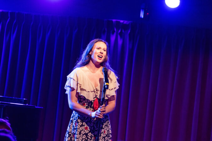 Rachel Handler is a 30 something white woman in a floral dress singing at a microphone stand. 