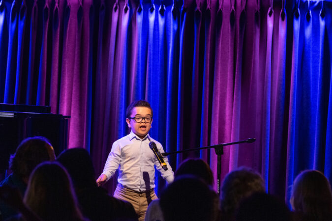 Nicolas Vivar is a teen Latin Little Person with glasses singing at a mic stand. He is wearing a light blue button down with khaki pants. 