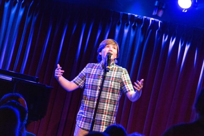 A white teenager in a plaid shirt  sings at mic stand. 